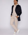 tall male model wearing beige long-inseam sweatpants, beige hoodie and dark navy mid-length coat, full-body lifestyle view