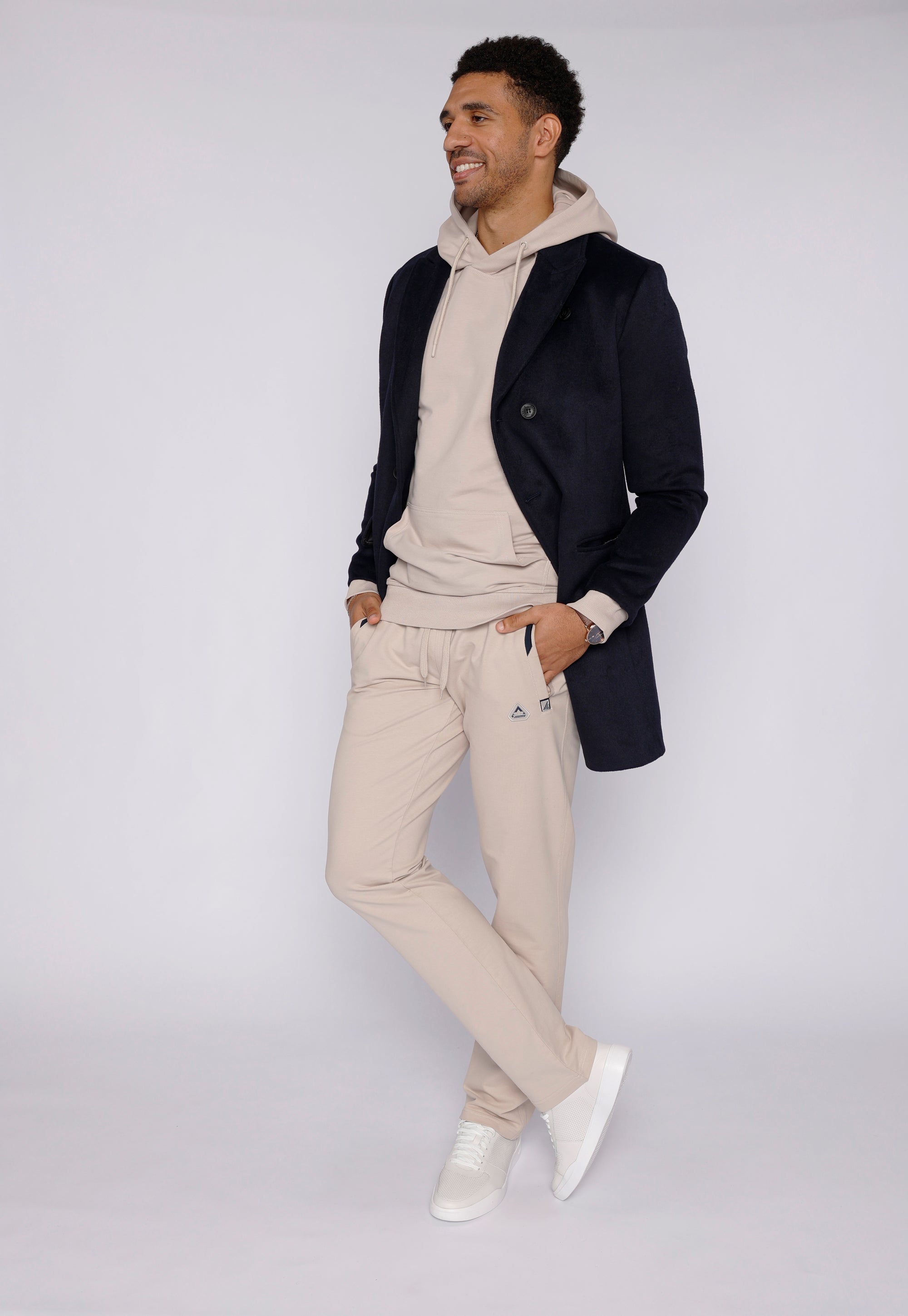 tall male model wearing beige long-inseam sweatpants, beige hoodie and dark navy mid-length coat, full-body lifestyle view