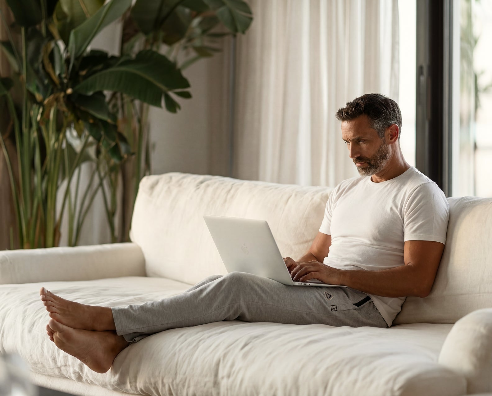 Man using a laptop on a white couch in a bright room with plants wearing white t-shirt and lounge pants