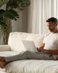 Man using a laptop on a white couch in a bright room with plants wearing white t-shirt and lounge pants