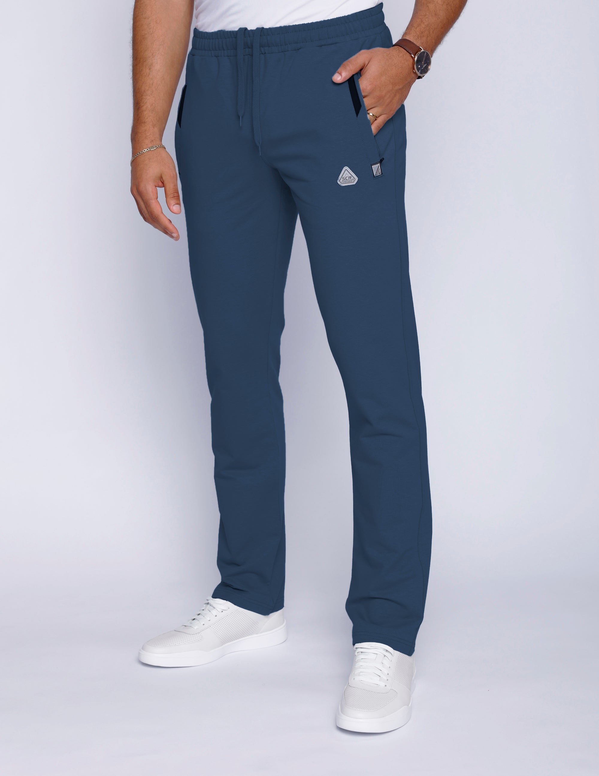 Front view of men’s steel blue sweatpants with straight leg cut, zippered pockets, and stretch waistband with drawcord, worn by male model in white t-shirt and white shoes.