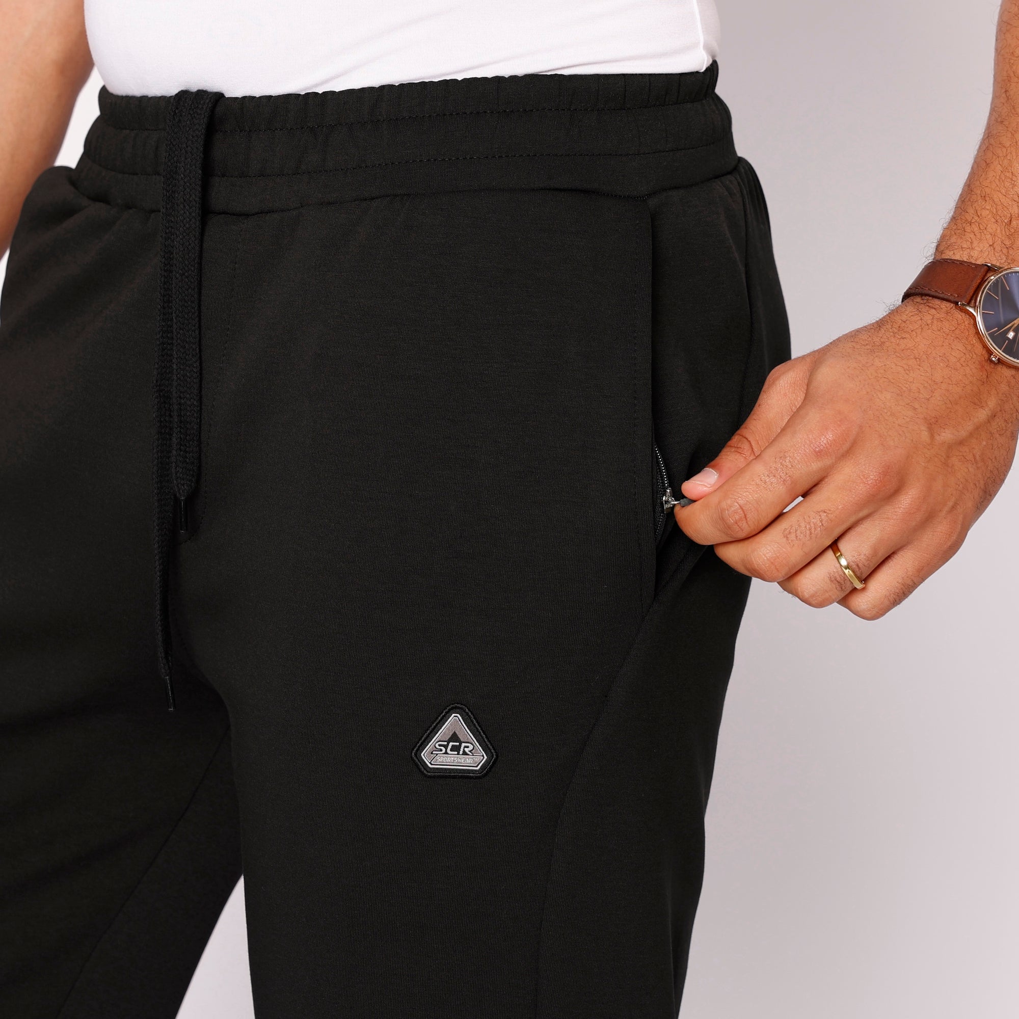 Close up of men's black straight leg athletic pants showing front zipper pocket, drawcord waistband, and SCR Sportswear logo.