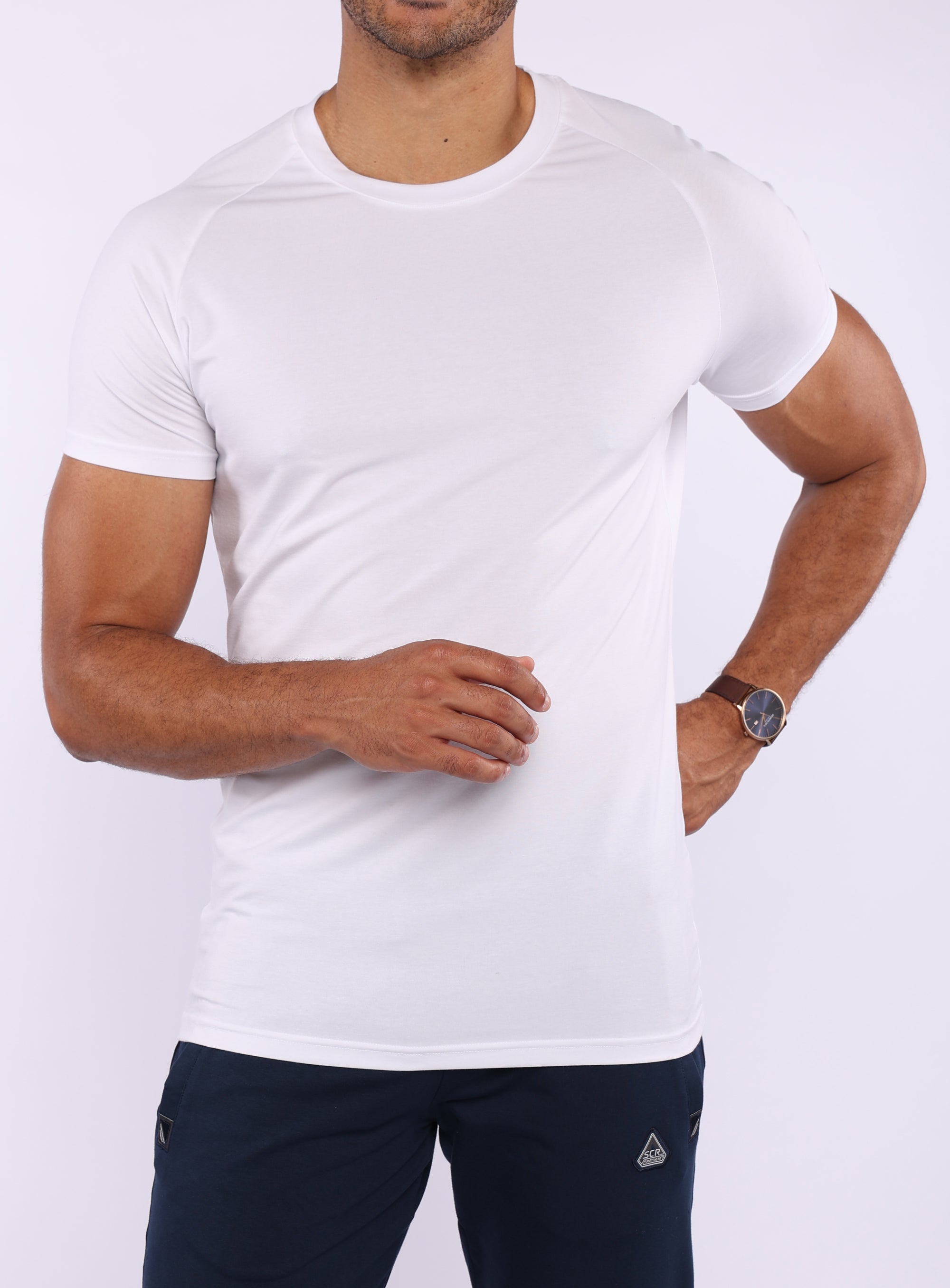A man wearing a white short-sleeve athletic crewneck t-shirt and dark navy pants, shown from the chin down against a light gray studio background.