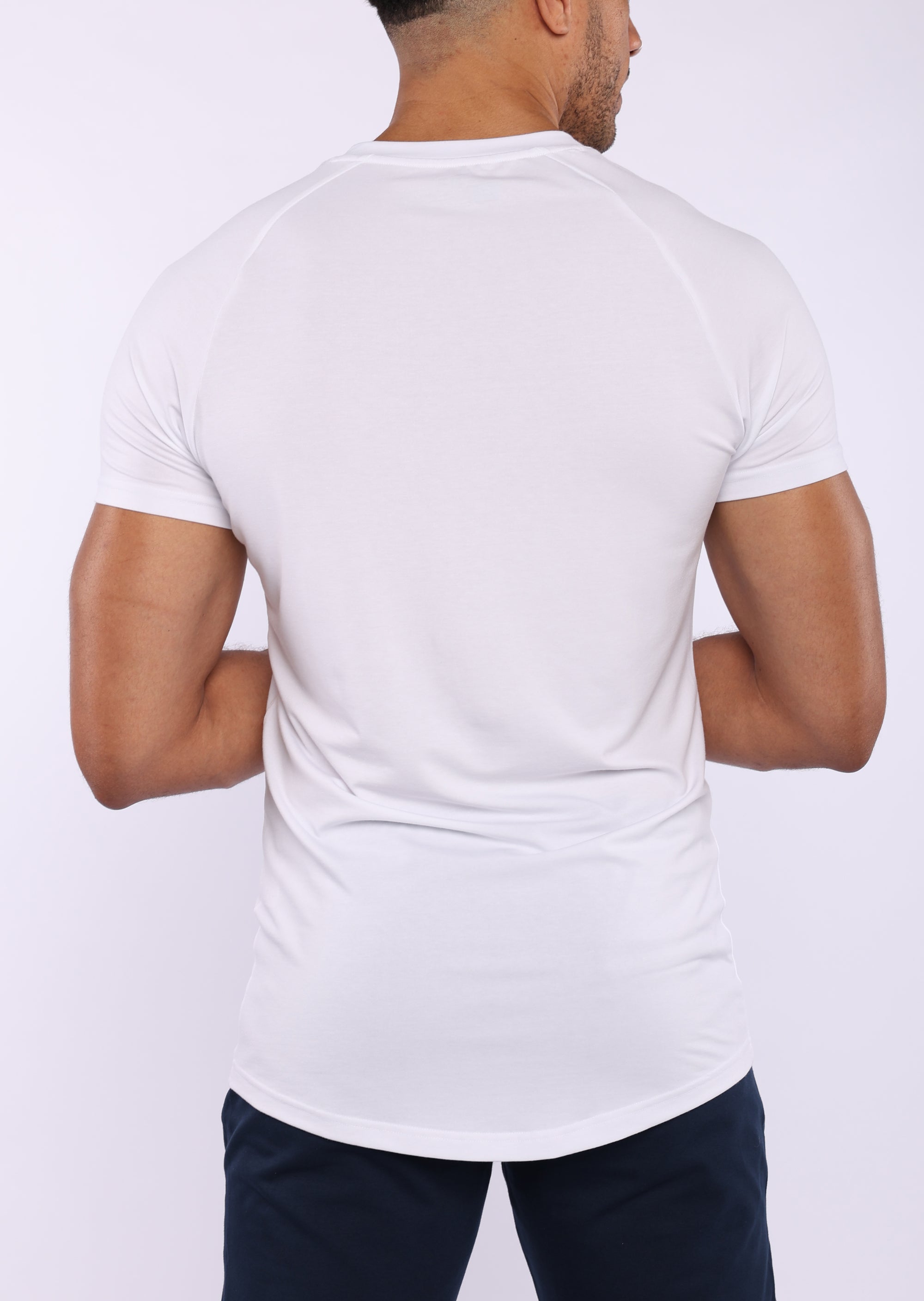 A man viewed from the back wearing a white short-sleeve athletic t-shirt and dark navy pants, set against a plain light gray studio background.