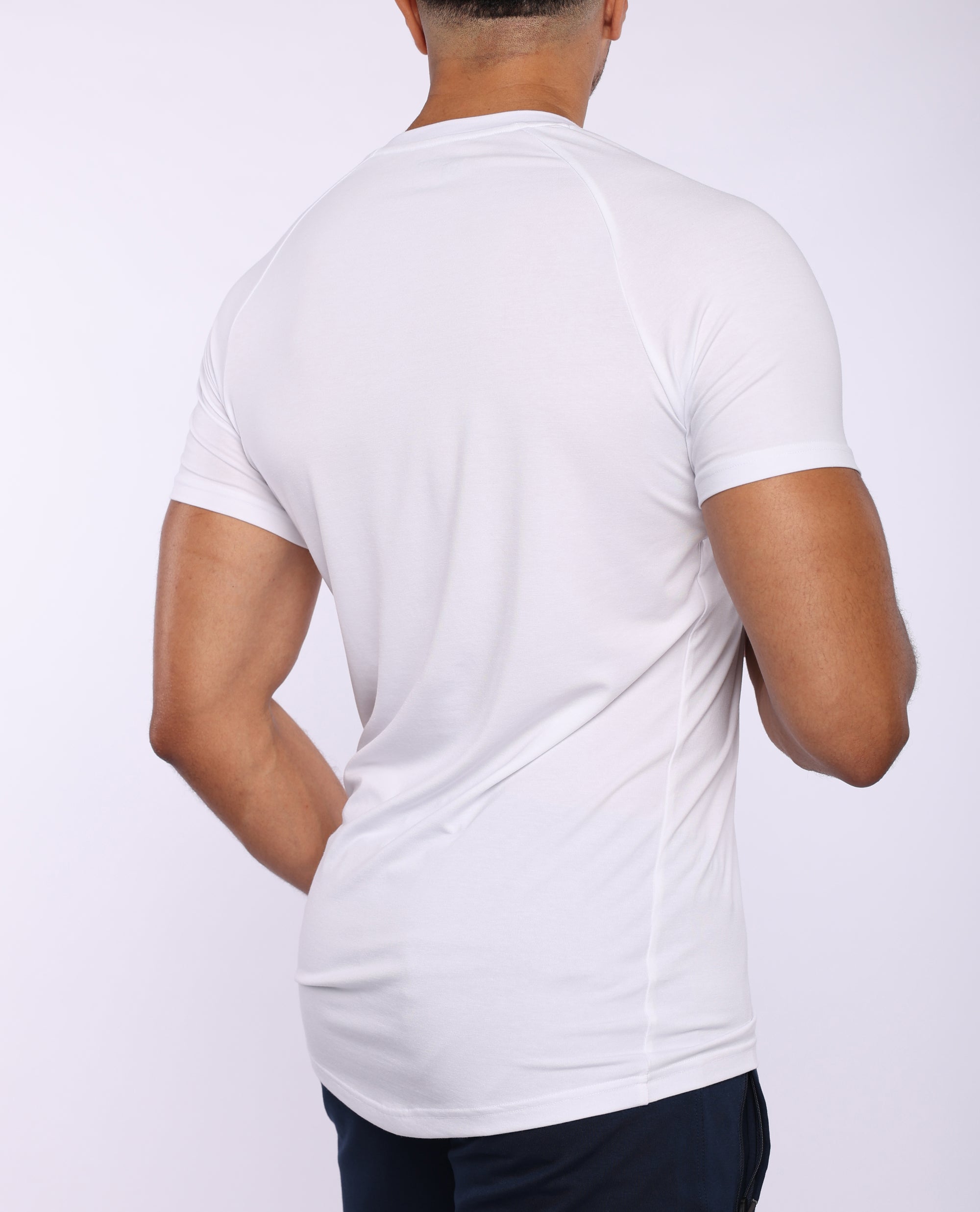 A man viewed from the back wearing a fitted white short-sleeve athletic t-shirt and dark navy pants, against a plain light gray studio background.