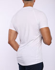 A man viewed from the back wearing a fitted white short-sleeve athletic t-shirt and dark navy pants, against a plain light gray studio background.