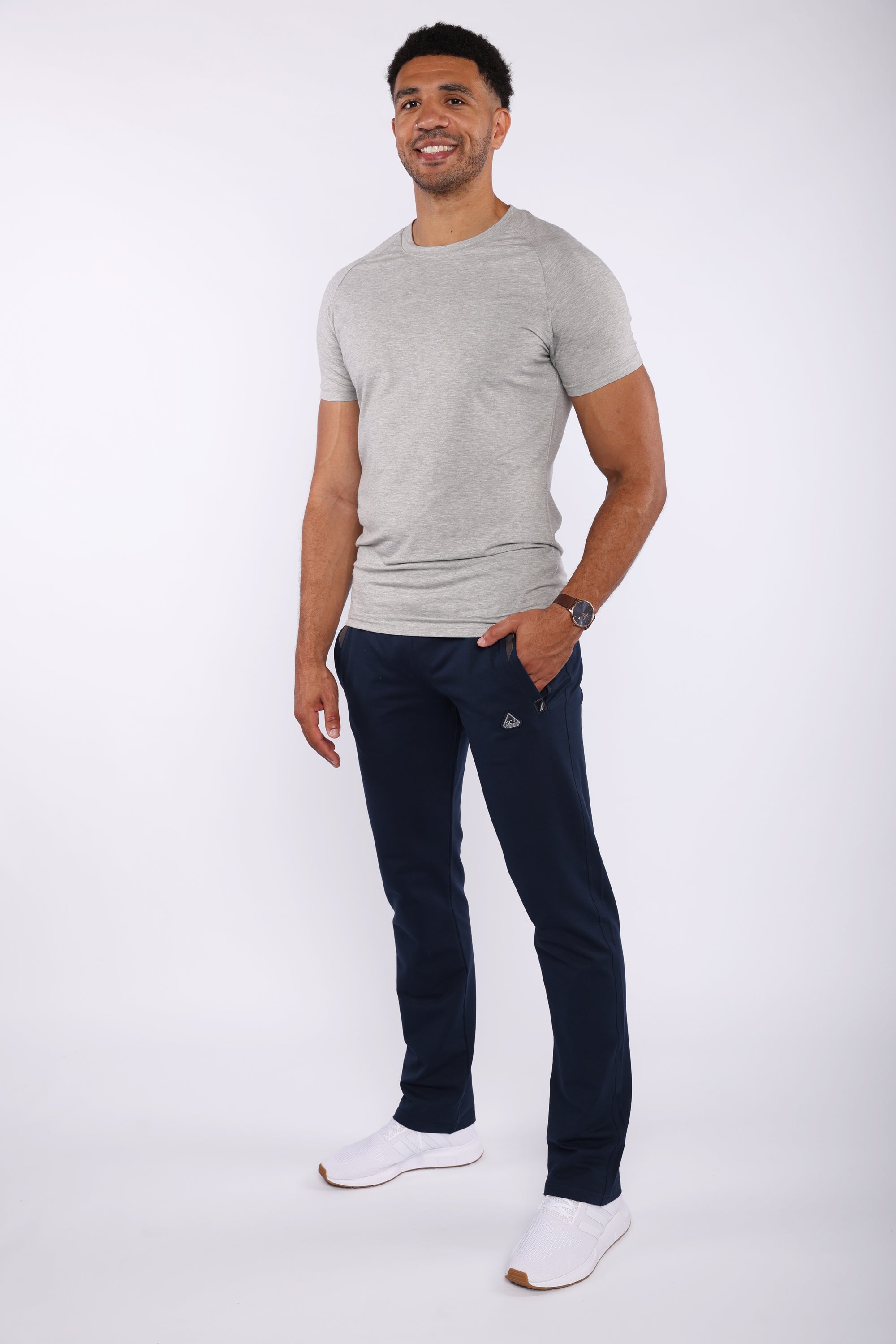 A man modeling a light gray crew-neck t-shirt and navy blue jogger pants with white sneakers, standing against a plain white studio background.