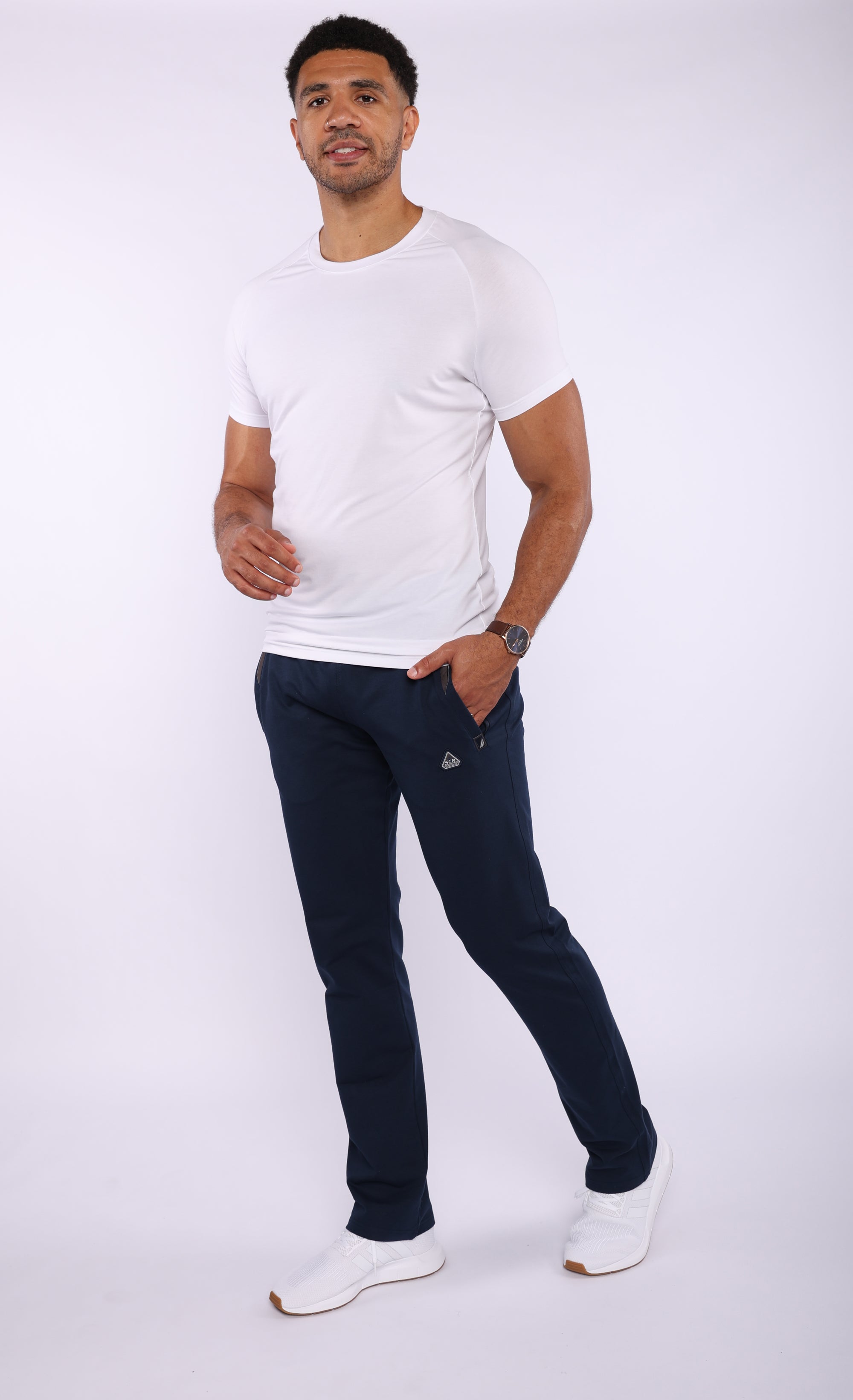 A man modeling a white crew-neck t-shirt and dark navy jogger pants with a small white embroidered logo on the thigh, paired with white sneakers, against a light gray studio background.