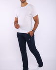 A man modeling a white crew-neck t-shirt and dark navy jogger pants with a small white embroidered logo on the thigh, paired with white sneakers, against a light gray studio background.