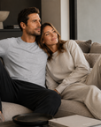 A man and a woman relax on a beige sofa in a modern living room. The man wears a light gray heather t-shirt and dark charcoal pants, while the woman wears a matching light beige lounge set.